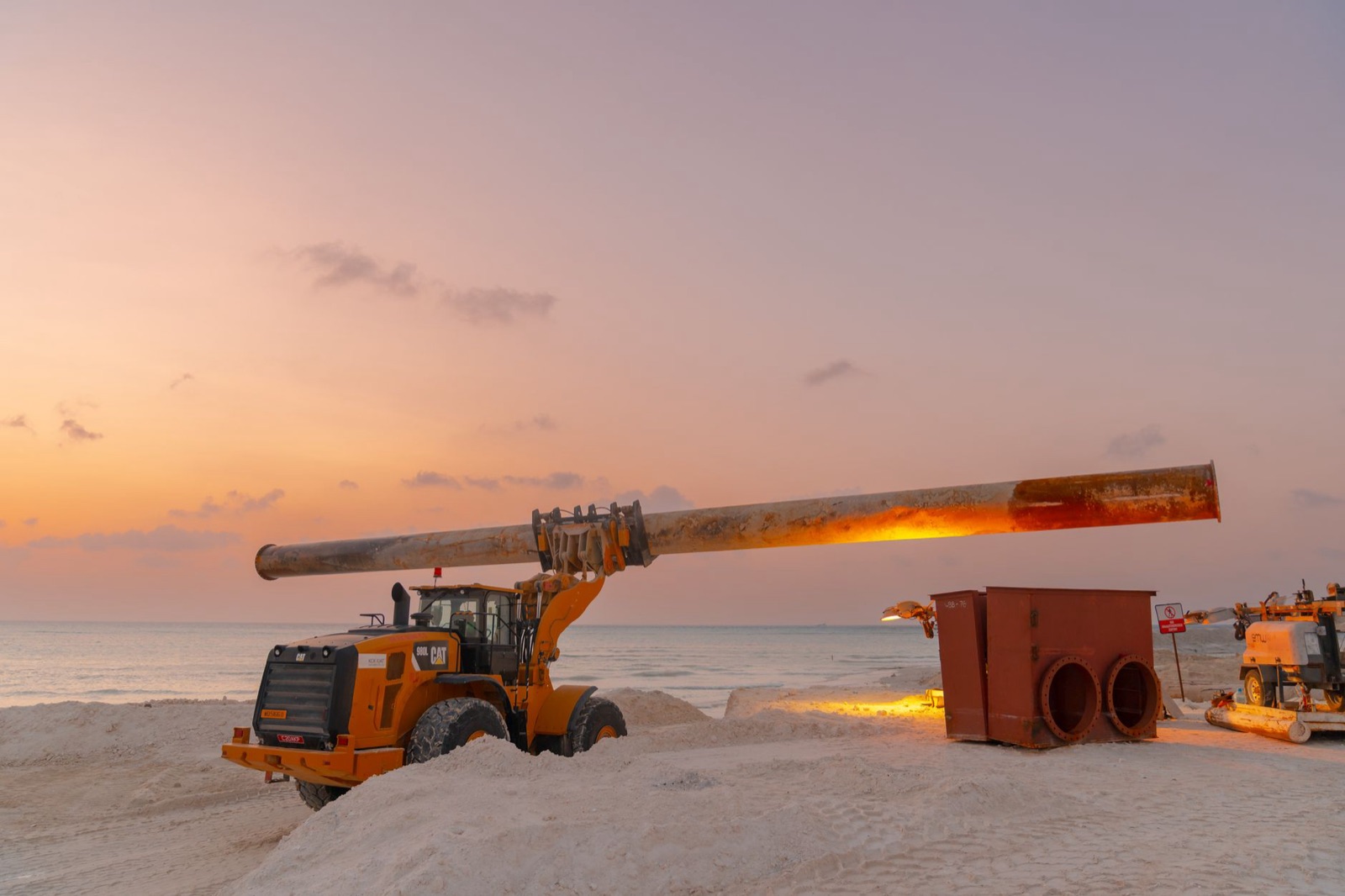 Loader carrying a long section of steel pipe at a coastal worksite at dusk