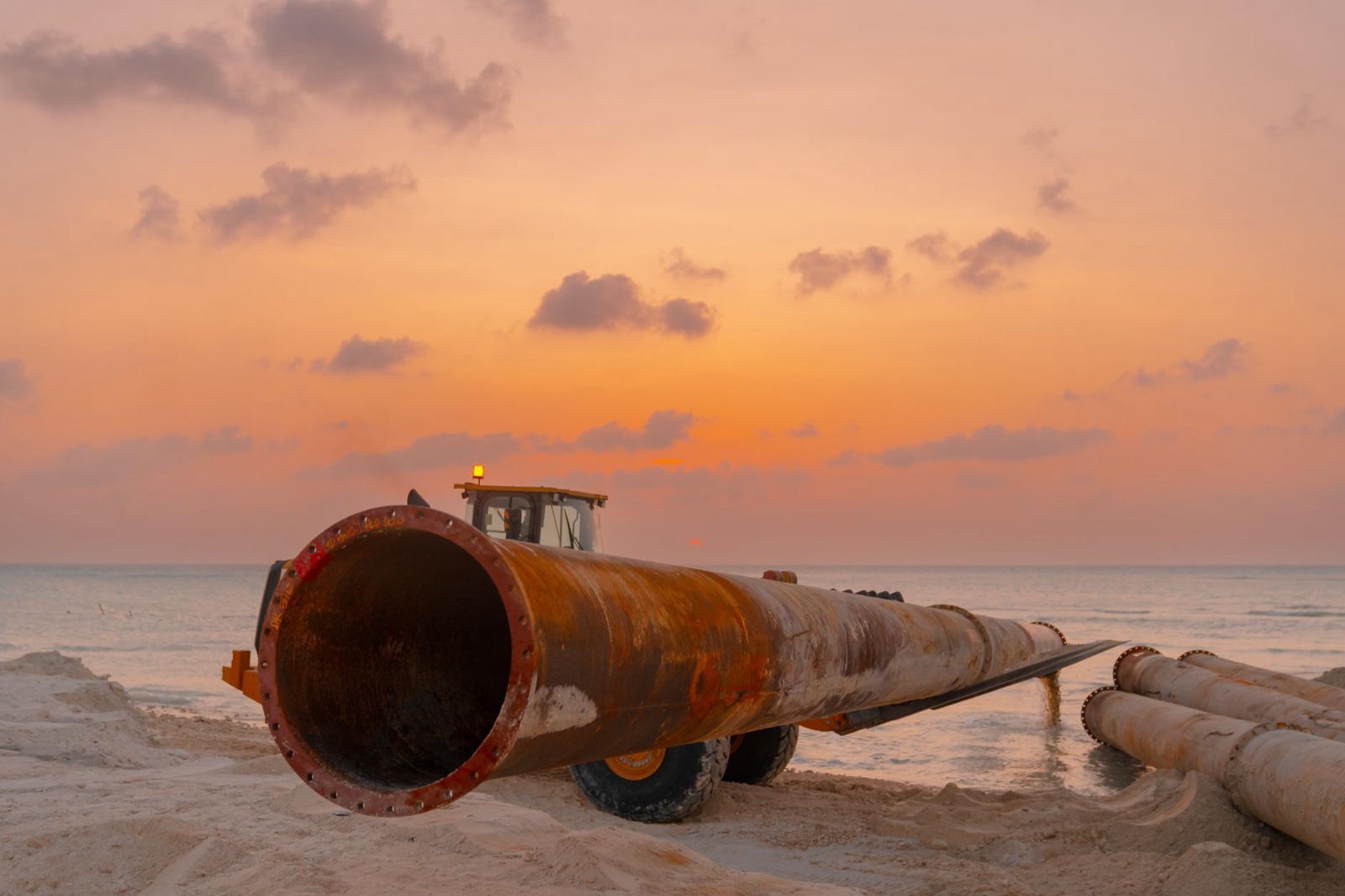 Steel pipe sections laid out beside a loader on a sandy pipeline worksite at sunset